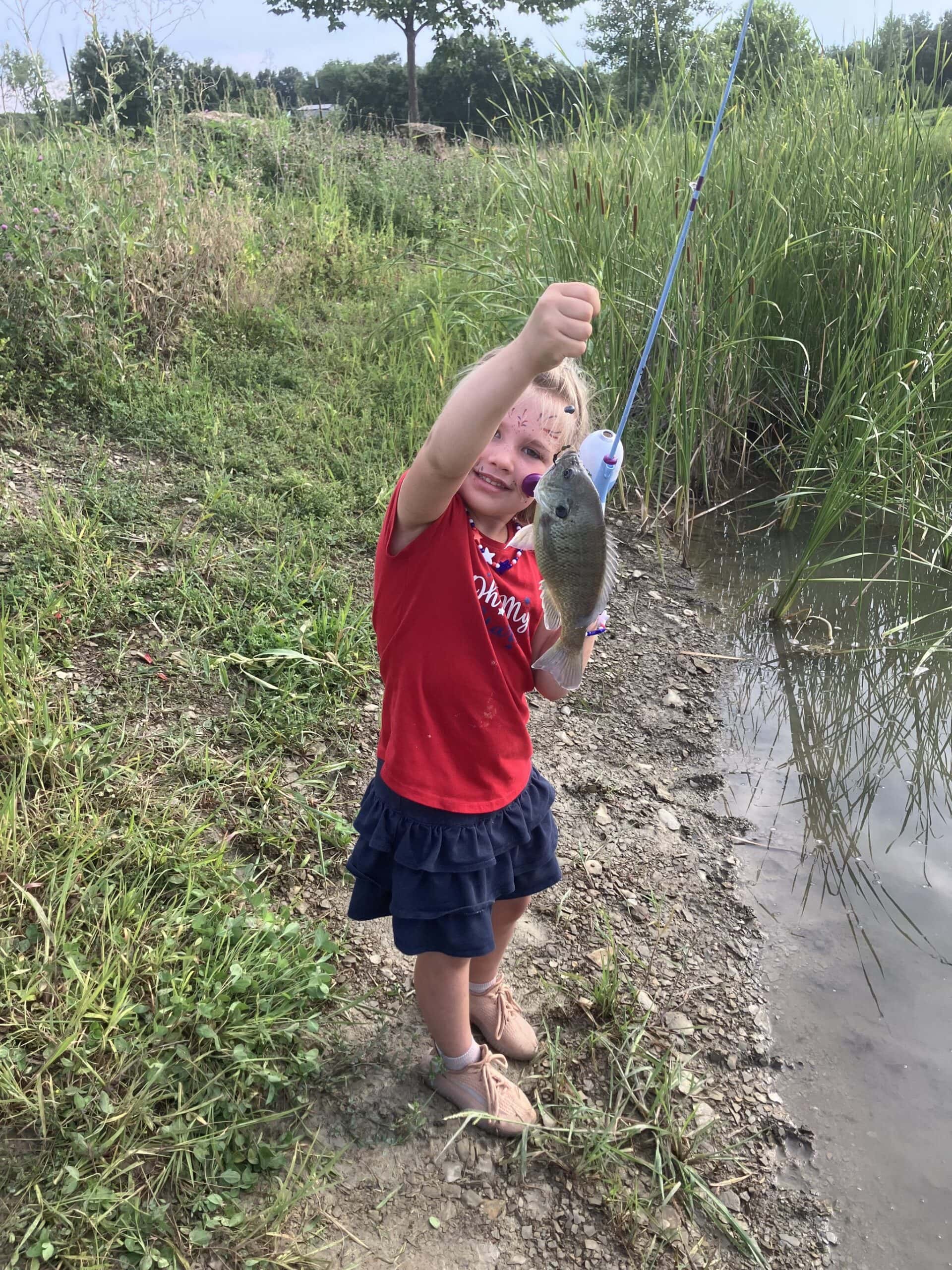 A young girl holding up a bluegill she caught, standing in the grass beside the cattails at the stocked pond at Selah Hills Ranch.
