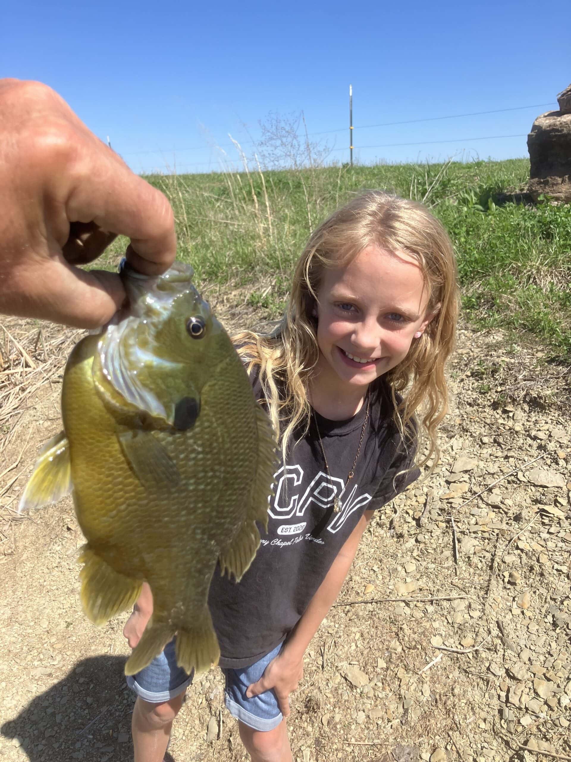 A young girl smiling beside a large bluegill held up on the line at the stocked pond at Selah Hills Ranch.