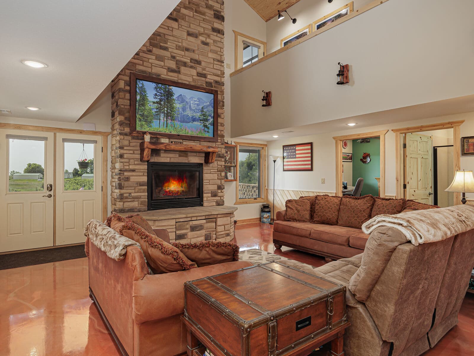 The great room at Selah Hills Ranch, a floor-to-ceiling stone chimney with a lit fireplace and live-edge mantel, deep sofas and a stained concrete floor under a vaulted ceiling.