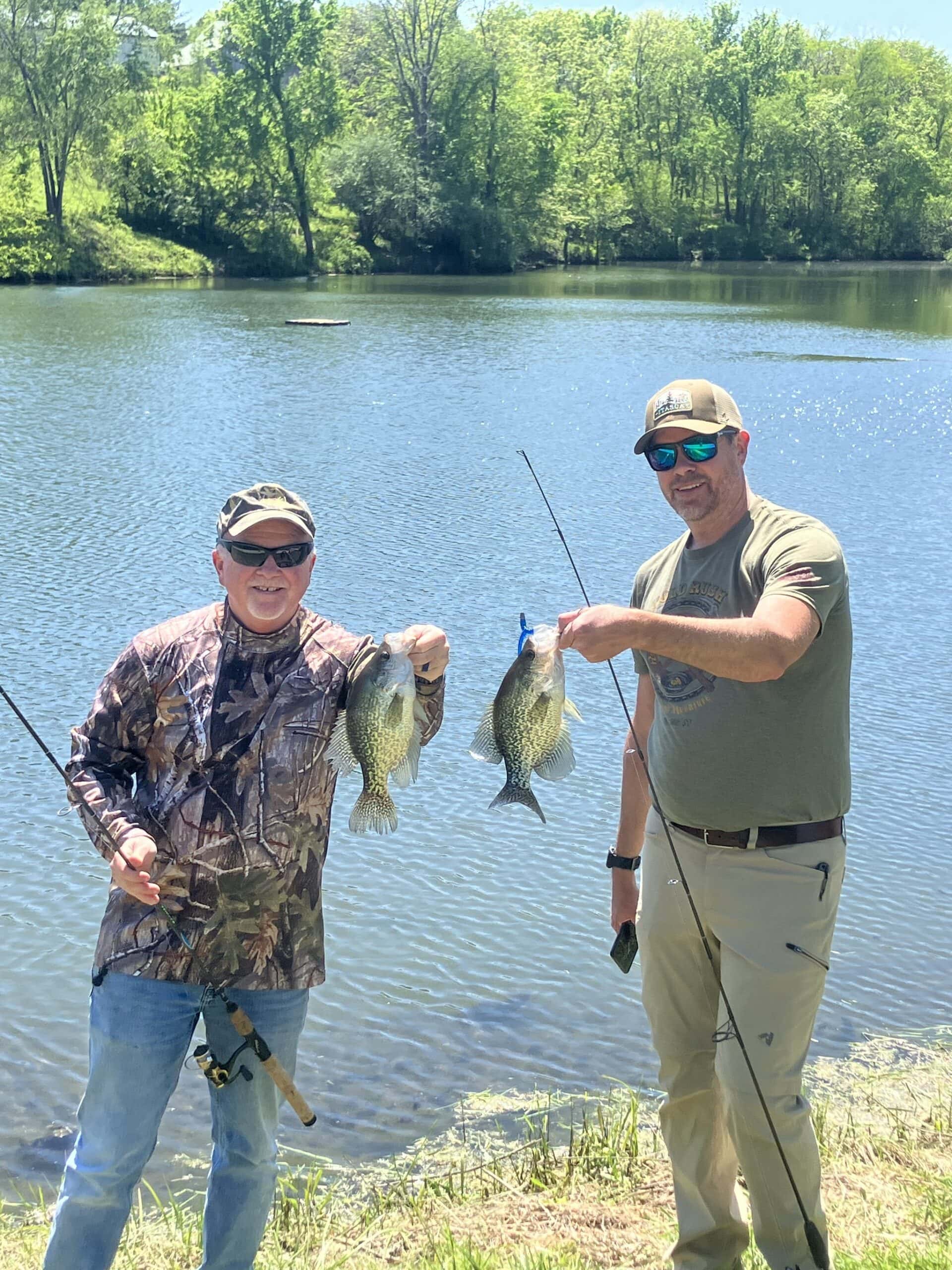Two guests holding up crappie they caught at the stocked pond at Selah Hills Ranch, with the water and the treeline behind them.