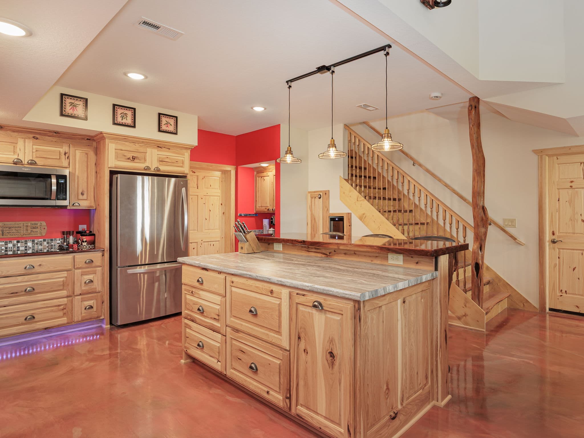 The guest kitchen at Selah Hills Ranch, hickory cabinets and a live-edge walnut breakfast bar beneath pendant lights, with the log staircase behind.
