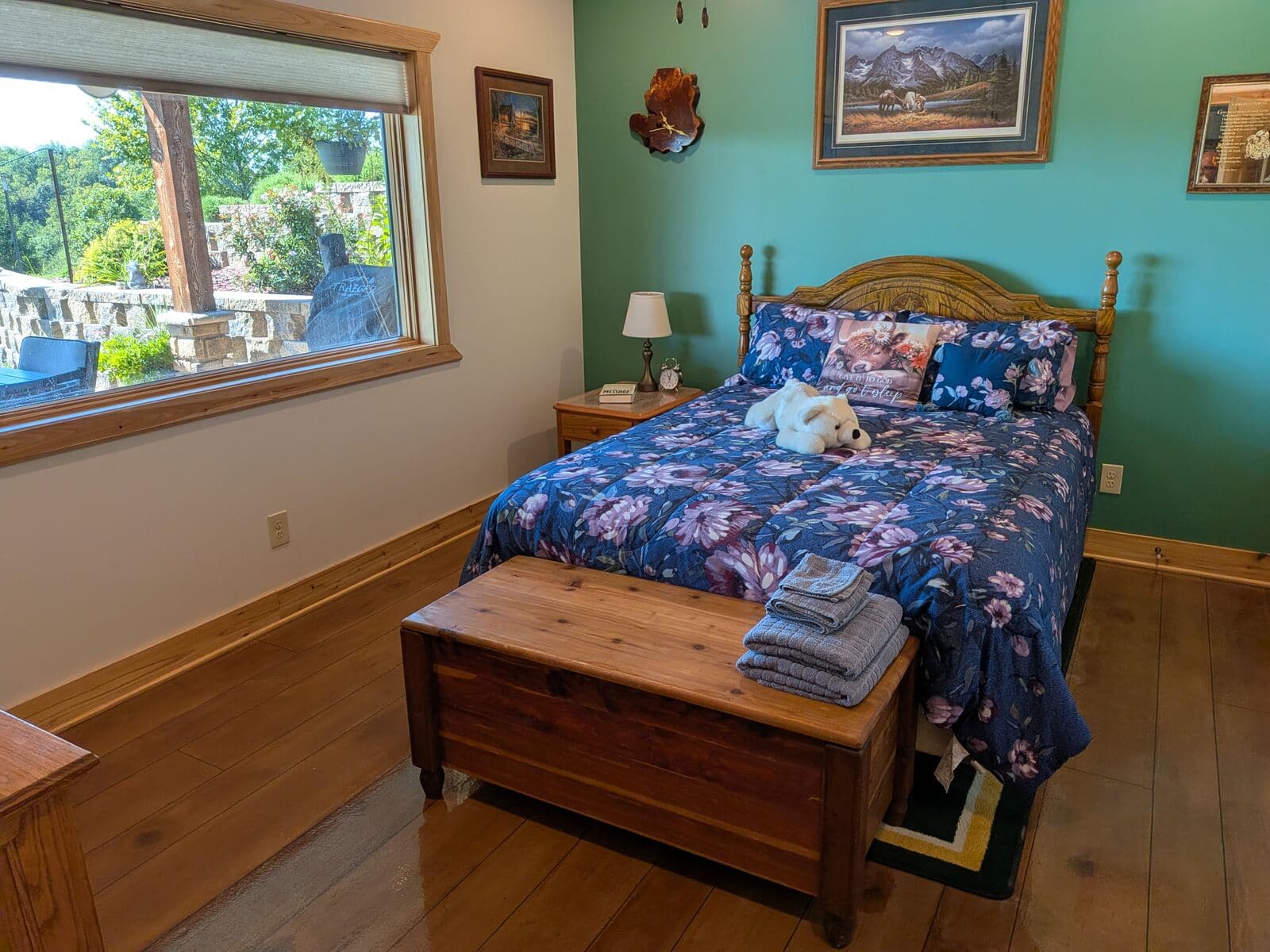The Queen Suite at Selah Hills Ranch, a queen bed with a carved oak headboard against a green wall, a cedar chest with folded towels at the foot of the bed, and a window looking out over the garden and stone wall.
