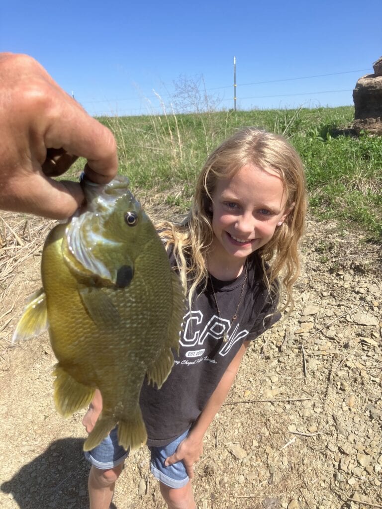 A young guest grinning on the bank at Selah Hills Ranch while a bluegill she caught is held up beside her.