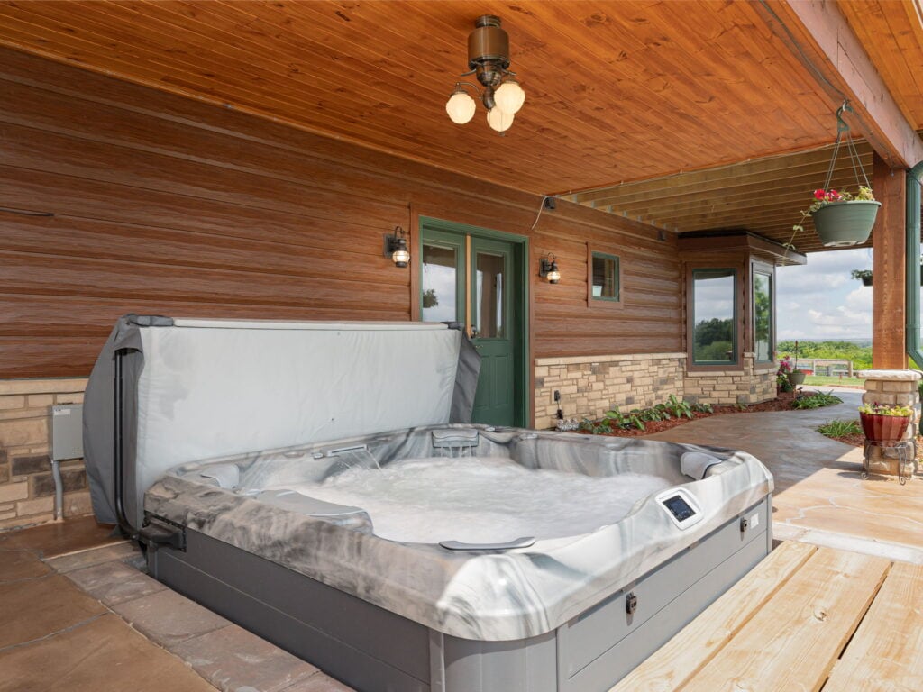 The eight person hot tub on the covered cedar patio at Selah Hills Ranch, lid open and water running, with the green door to the guest level and the valley beyond.